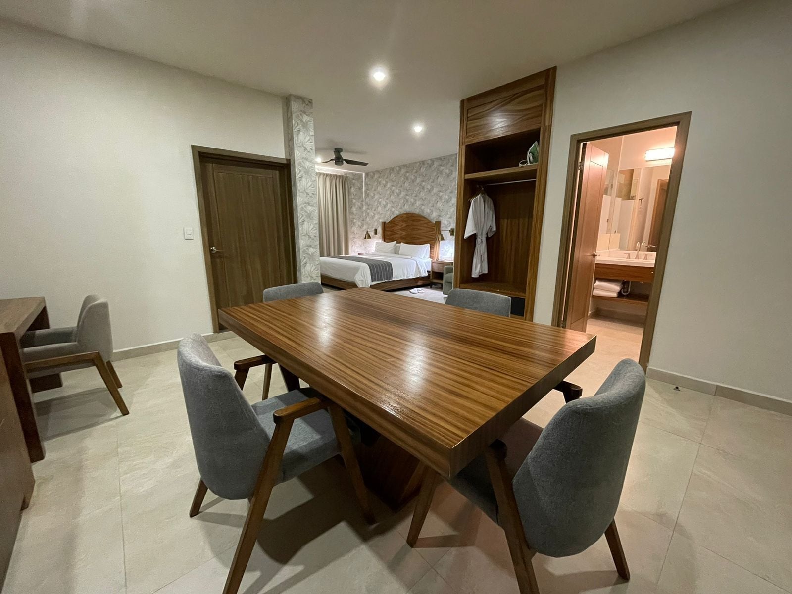 Uma sala de jantar com mesa de madeira e quatro cadeiras, uma cama com cabeceira, um guarda-roupa e um banheiro visível por uma porta aberta.