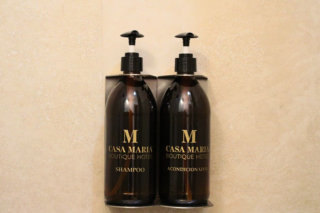 Dos botellas marrones oscuro con dosificador, etiquetadas 'Casa Maria Boutique Hotel Champú' y 'Casa Maria Boutique Hotel Acondicionador'.