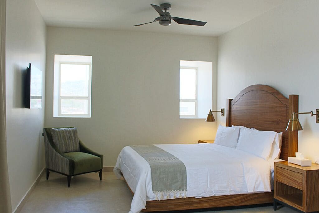 Quarto com cama, cadeira, mesa de cabeceira, ventilador de teto e duas janelas.