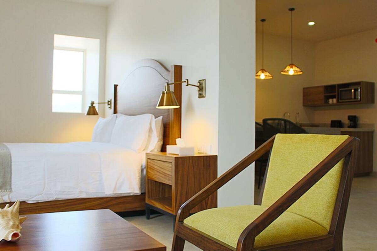 Une chambre d’hôtel comprenant un lit, une table de chevet, une lampe, une chaise et donnant sur la cuisine.
