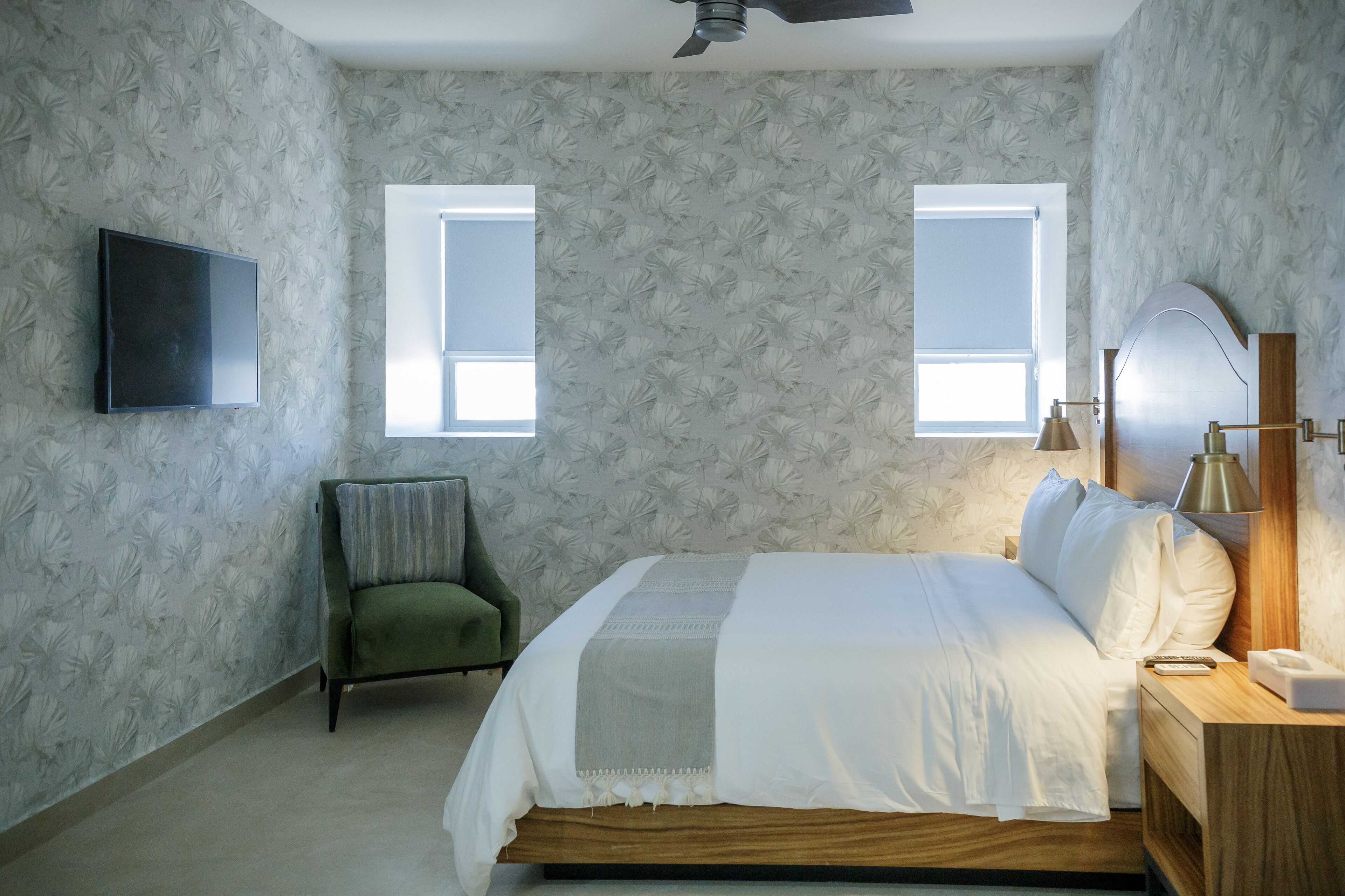 Un dormitorio con una cama, dos ventanas, un televisor, una silla y una mesita de noche con una lámpara.