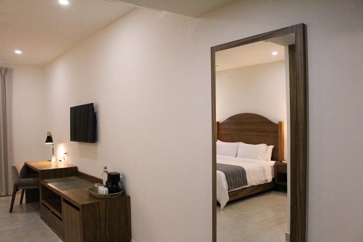 A hotel room with a wooden desk, a chair, a flat-screen TV, a bed with white bedding, and a large mirror.