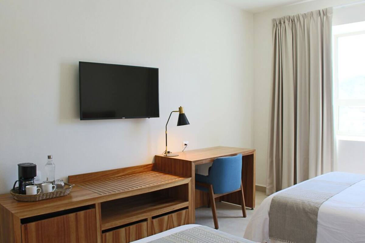Une chambre d’hôtel avec un bureau en bois, une chaise, une télévision, un lit et une fenêtre avec des rideaux.