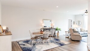 A modern living room with a dining area, featuring a wooden dining table, white chairs, a sofa, and a coffee table. - Stoney Ridge · Cozy 1st Level King Bed Suite - 2 bed 2 bath! (Forest)
