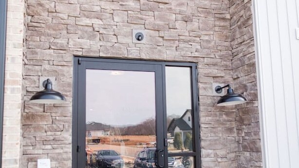 A building entrance with a stone facade, a glass door, and two wall-mounted lights. - Stoney Ridge · Cozy 1st Level King Bed Suite - 2 bed 2 bath! (Forest)