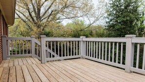 A wooden deck with a white railing, surrounded by trees and a brick building. - Charming Single Level Home • Convenient 3 Bedroom (Lynchburg)