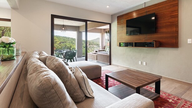 A modern living room with a large sectional sofa, a flat-screen TV mounted on a wooden wall, and a dining area visible through a glass door. - Moderna Villa con Hermosa Vista al Lago (Valle de Bravo)