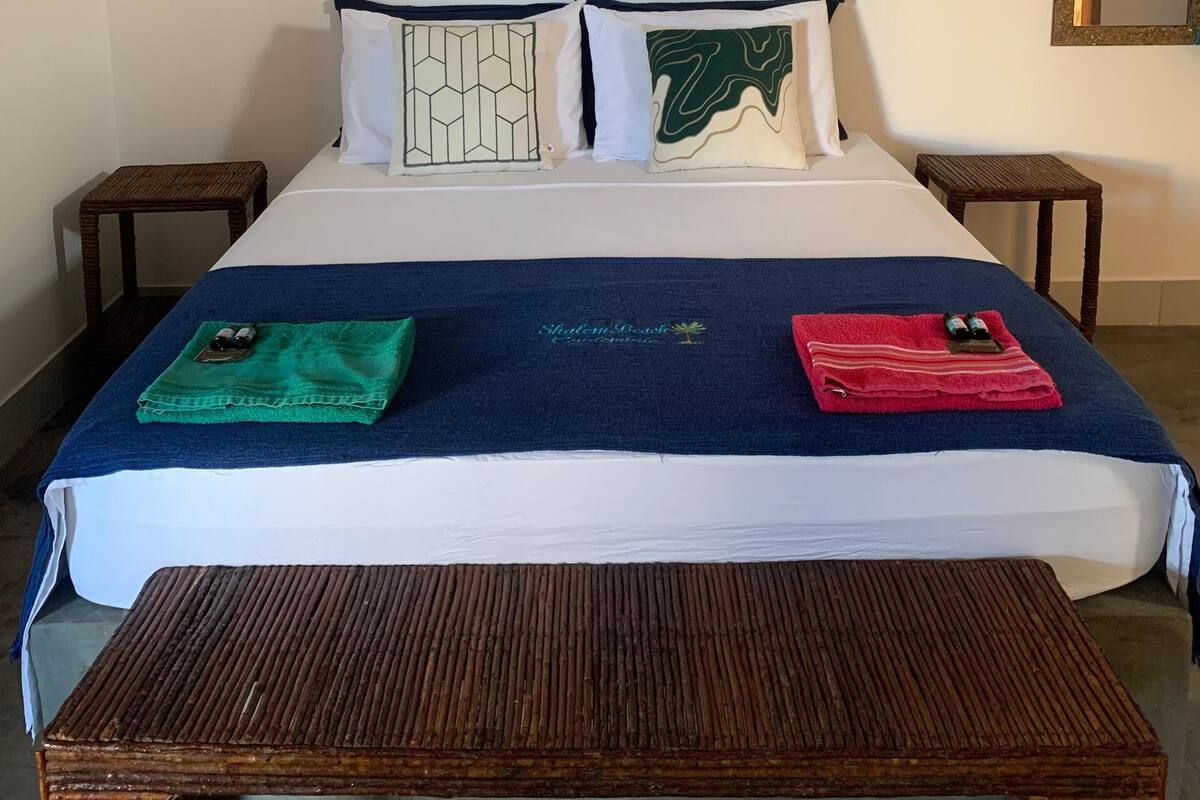 Un lit bien fait, recouvert d’une couvre-lit bleue et blanche, avec deux oreillers aux motifs géométriques, une tête de lit en osier et une table de chevet en bois.