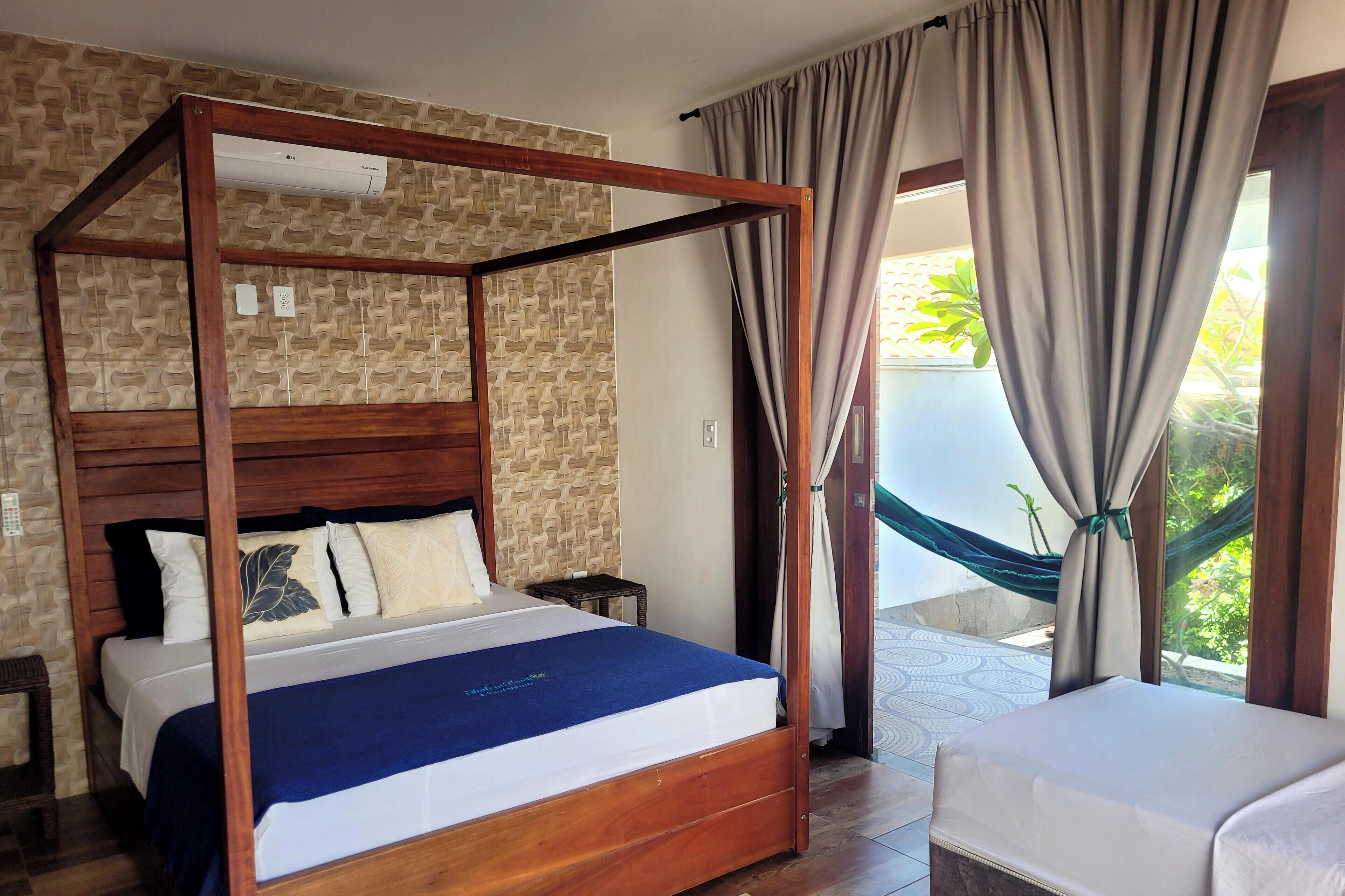 A four-poster bed with a wooden frame, a brick-patterned wall, and a view of a garden through a window with curtains.