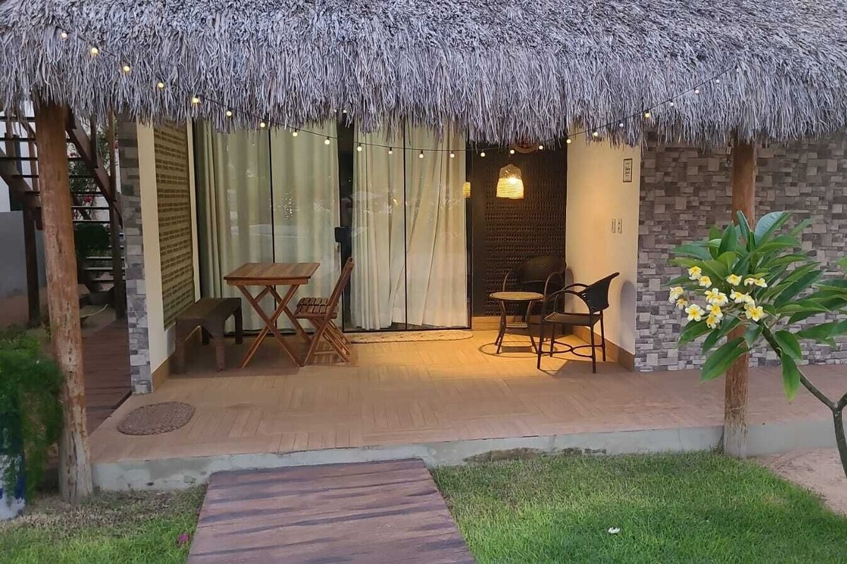 Ein Sitzbereich im Freien mit Reetdach, einem Tisch und Stühlen, ein kleiner Garten mit Baum und Blumen, und ein Weg zum Hauseingang.
