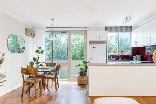 A modern kitchen with white cabinets, a dining area with wooden chairs, and a view of greenery outside.