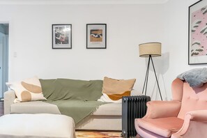 A living room with a sofa, armchair, floor lamp, radiator, and two framed pictures on the wall.