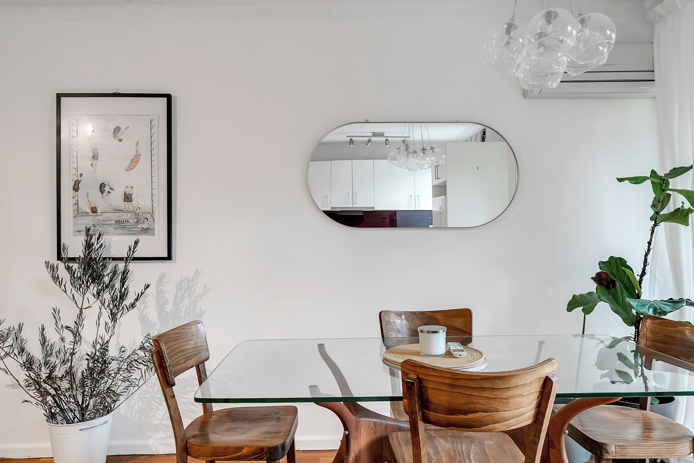A dining area with a glass table, wooden chairs, a framed artwork, a mirror, and a potted plant.