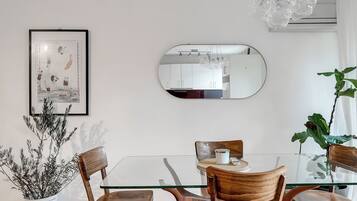 A dining area with a glass table, wooden chairs, a framed artwork, a mirror, and a potted plant.