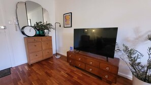 A living room with a wooden dresser, a large flat-screen TV, a clock, a potted plant, and a framed picture on the wall. - Cosy Bright 1 BDR Apt Lane Cove (Lane Cove)