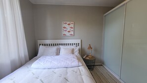 A neatly made bed with a white headboard and pillows, a bedside table with a lamp, and a wall with a framed picture.