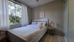 Een slaapkamer met een bed, een nachtkastje met een lamp, een raam met gordijnen en een muur met een flamingo schilderij.