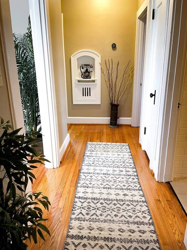 A hallway with wooden flooring, a patterned rug, a white wall-mounted shelf with a decorative item, and a potted plant. - 12. Historic " Turret House" established in 1937 (Boaz)