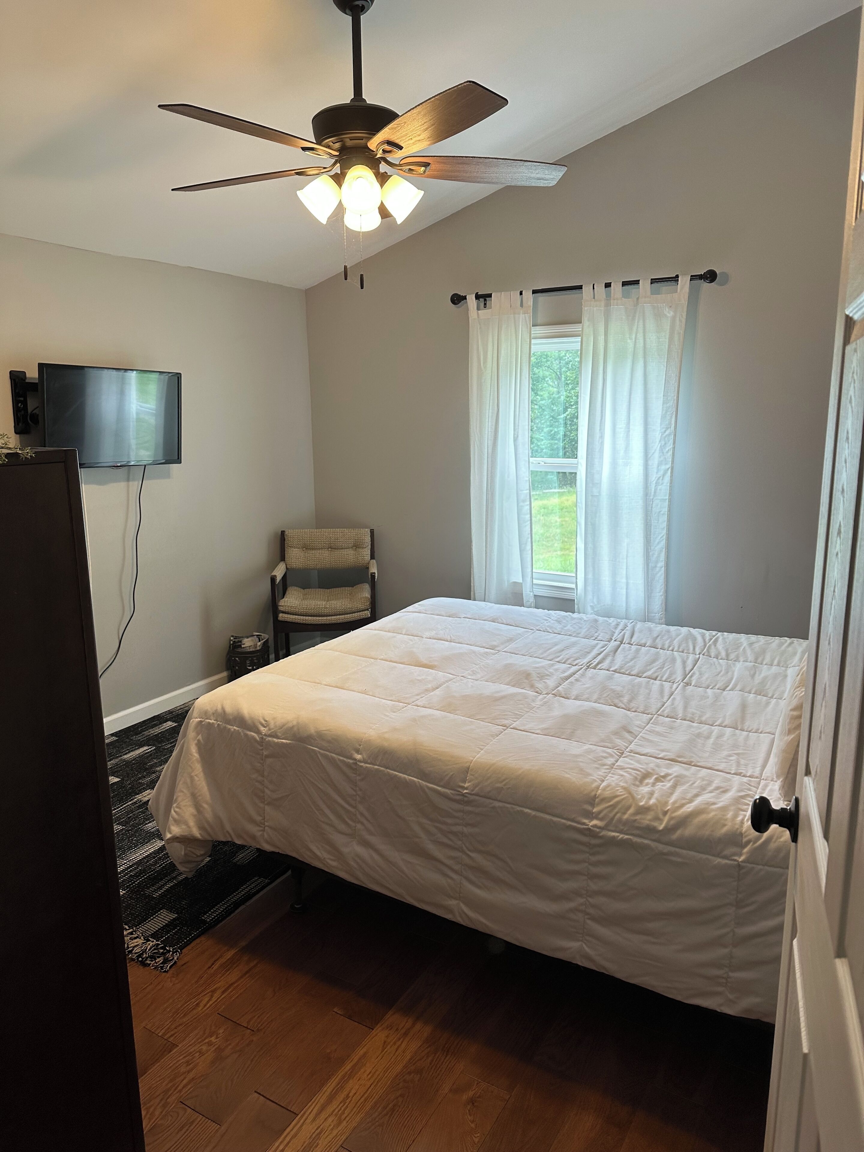 A bedroom with a bed, a ceiling fan, a television, and a window with curtains.