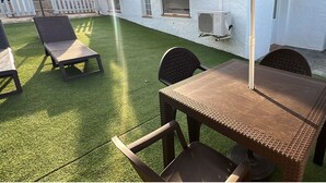 Un espace extérieur aménagé en terrasse, avec une table et des chaises, un bâtiment blanc en arrière-plan et une clôture blanche en bois.