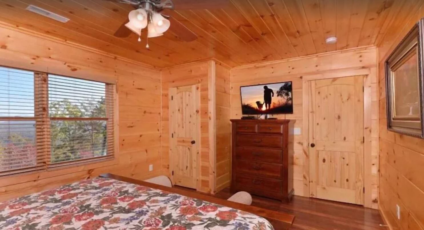 A wooden cabin room with a bed, a dresser, a television, and a window with blinds.