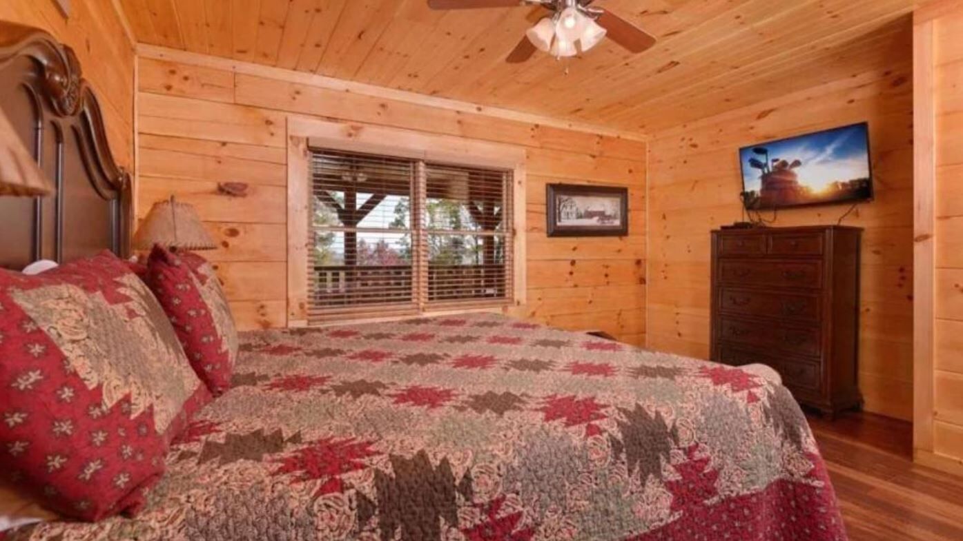 A cozy bedroom with a bed, wooden walls, a window with blinds, a dresser, and a television on the wall.