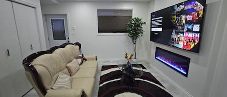 A living room with a beige sofa, a TV mounted on the wall, a small table with a vase, and a decorative rug.
