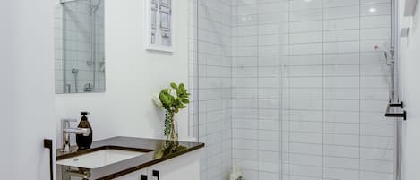 A modern bathroom with a white sink, mirror, and toilet, a shower with glass doors, and wooden flooring.
