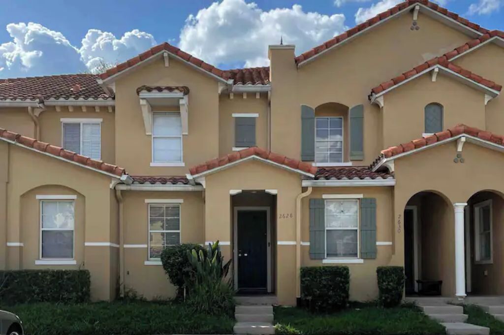A two-story house with a beige facade, red-tiled roof, and a front entrance.