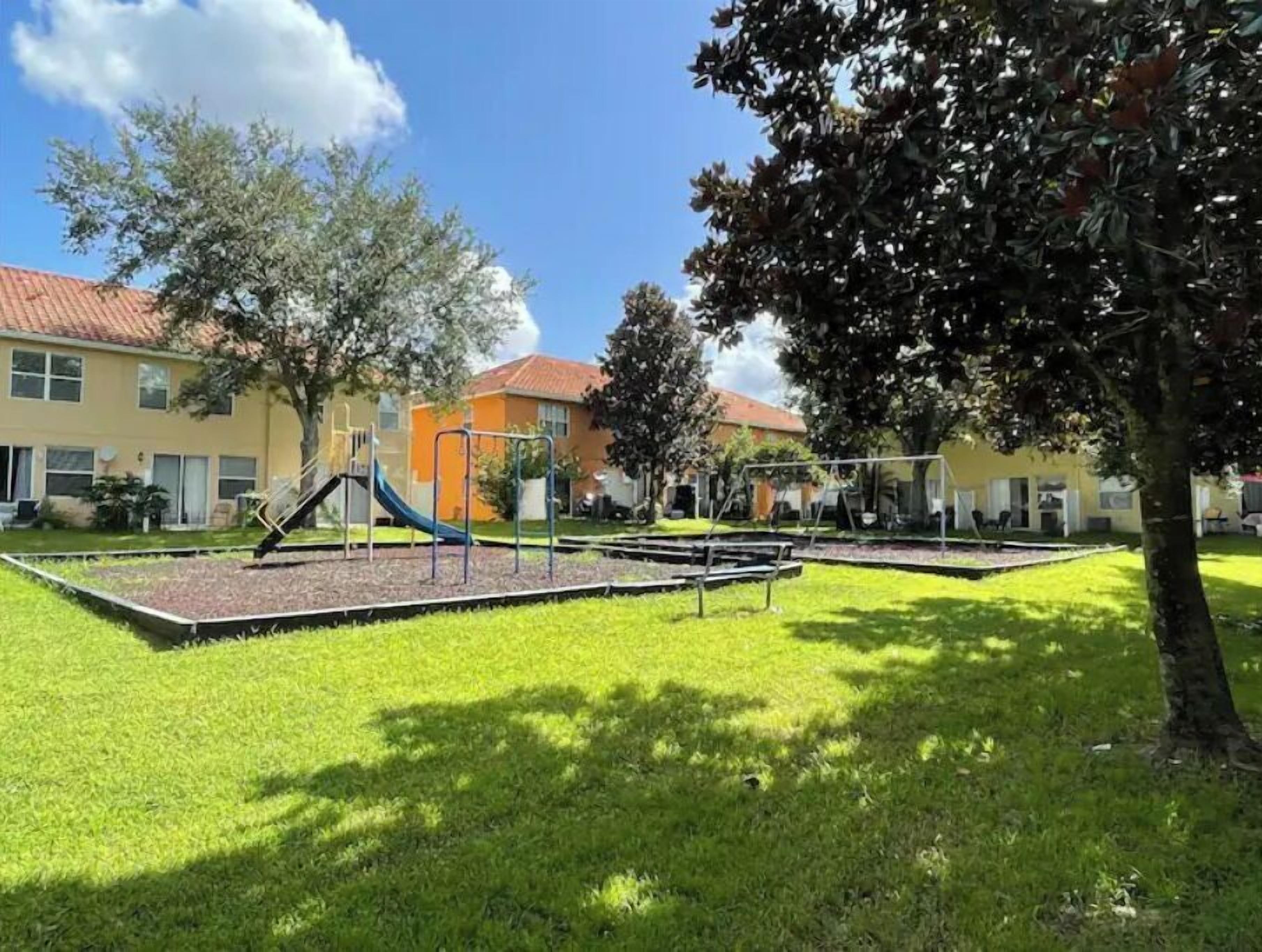 A playground with slides and swings, surrounded by grass and trees, with buildings in the background.