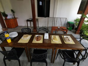 A dining area with a wooden table set for four, surrounded by black chairs. There are plates of food on the table, a bottle of orange juice, and a water dispenser. The setting includes a concrete floor, a wooden pillar, and a window with a black frame. - Palm Gate Maldives (Thulusdhoo Island)