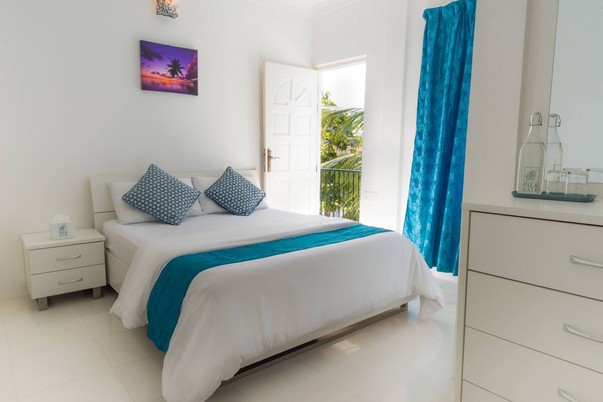 Un dormitorio con cama, mesita de noche, cómoda, ventana con vista a follaje y un cuadro en la pared.