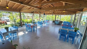 A covered outdoor seating area with blue chairs and tables, surrounded by glass walls with a view of greenery and a parked car.