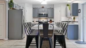 A modern kitchen with grey cabinets, a stainless steel refrigerator, a microwave, and a dining area with a long table and black chairs. - Poolside Cocoa Oasis with Tiki Bar (Port Saint John)