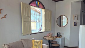 Una habitación con sofá, puf, ventilador de techo, ventana con contraventanas abiertas, una mesita con cafetera y un espejo en la pared.