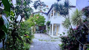 Un estrecho sendero adoquinado que conduce a un edificio blanco de estilo arquitectónico tradicional, rodeado de exuberante vegetación y plantas con flores.