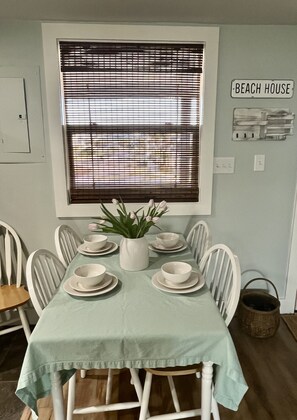 A dining area with a table set for four, white chairs, a potted plant, and a sign reading "Beach House" on the wall.