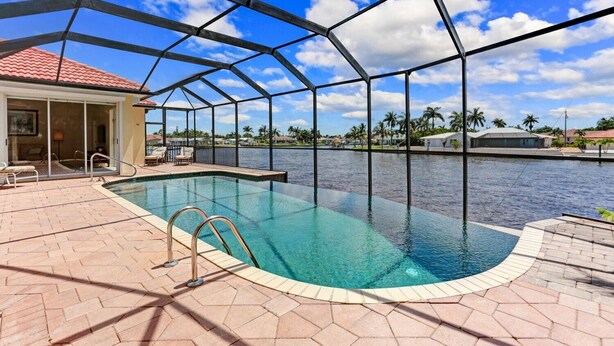 Una piscina con cerramiento de vidrio, un patio cubierto y vistas a un cuerpo de agua con palmeras al fondo.