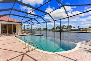 A pool with a glass enclosure, a covered patio area, and a view of a water body with palm trees in the background. - The Hammock - Beautiful Gulf access - 4 bedroom home - with heated pool (Cape Coral)