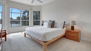 Un dormitorio con una cama grande, un ventilador de techo, una mesita de noche y vistas al exterior a través de amplias ventanas.