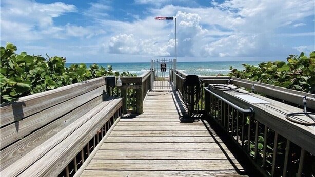 Une promenade en bois menant à une plage, avec un mât de drapeau et une porte.