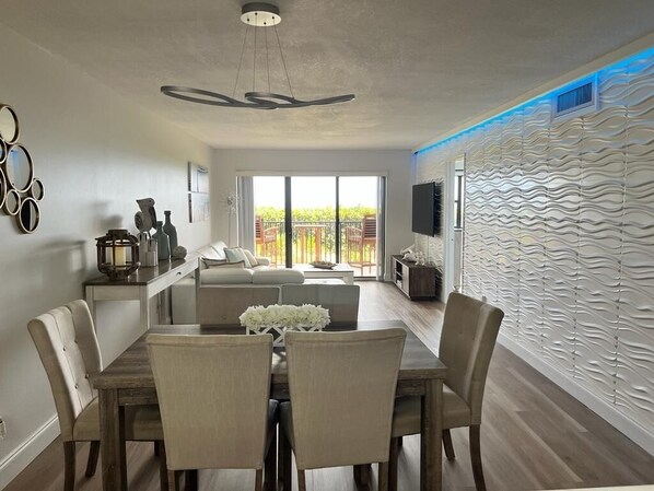 A modern living room with a dining area, a sofa, a TV mounted on the wall, and a balcony with a view. - Beach Condo (Jensen Beach)