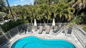 Une piscine entourée d’une clôture blanche et de palmiers, avec des chaises longues et des parasols.