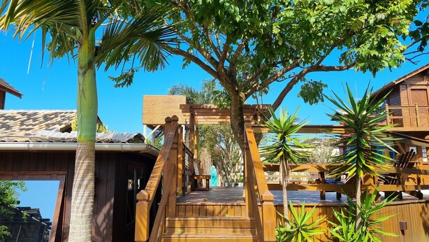 A wooden deck with a staircase leading to a covered area, surrounded by tropical plants and a clear blue sky. - Arte da Tribo Hsotel & Camping - Hostel (Imbituba)