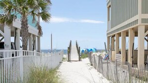 A sandy pathway leading to a beach with a white fence on one side and a building with a balcony on the other.