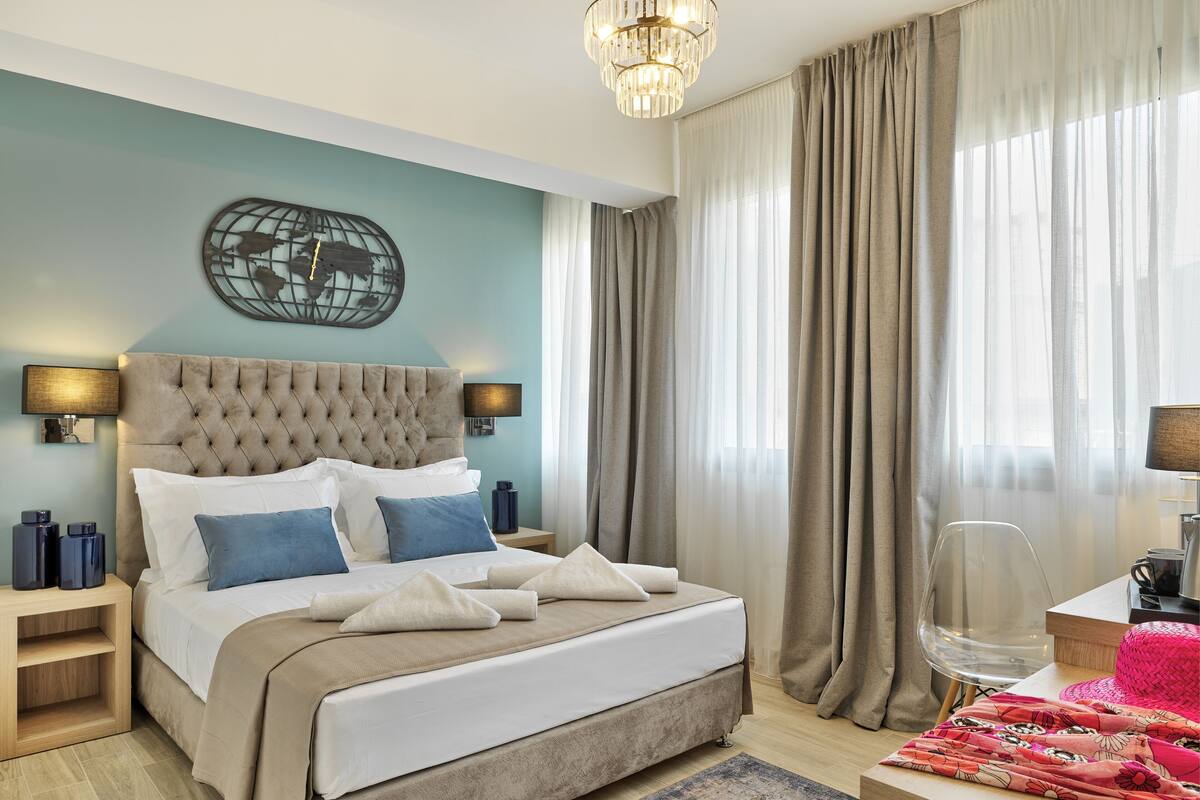 A modern hotel room with a large bed, a chandelier, and a view of the city through sheer curtains.