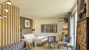 A modern hotel room with a large bed, a grey sofa, two armchairs, a small table with a vase, and a wooden cabinet with glass doors. - ACROPOLIS CITY LIFE BH (Athens)