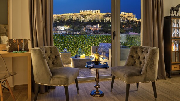 A room with a view of a historic building, two armchairs, a small table with a candle, and a cabinet with bottles. - ACROPOLIS CITY LIFE BH (Athens)