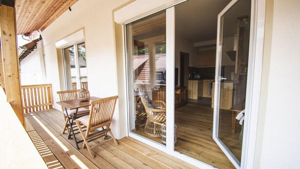 Eine Holzterrasse mit Tisch und Stühlen, eine Glasschiebetür, die zu einer Küche führt, und eine Holztreppe.