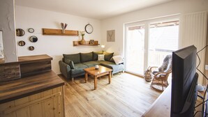 Ein gemütliches Wohnzimmer mit einem Couchtisch aus Holz, einer Couch und einem Fernseher. Regale mit dekorativen Gegenständen und ein großes Fenster für natürliches Licht.